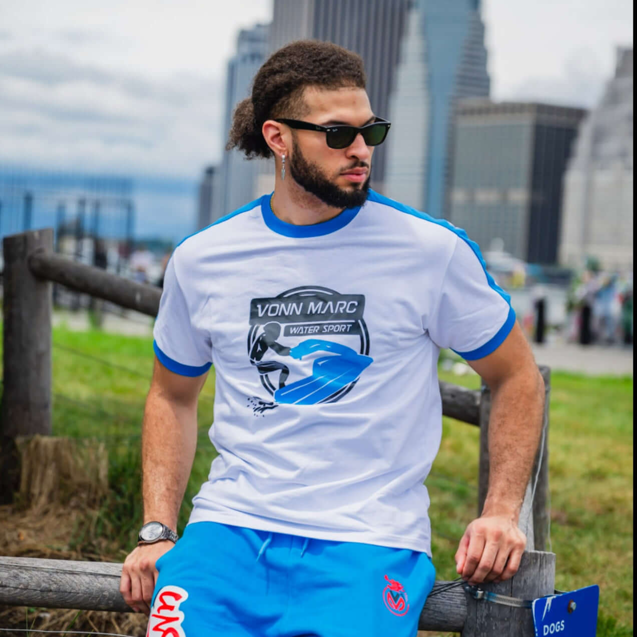 Man wearing a white t-shirt with blue accents and blue pants, standing outdoors with cityscape in the background.